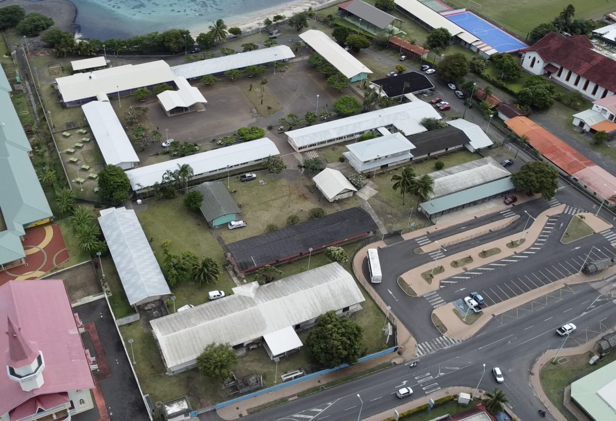 Le collège de Papara vu du ciel, à la verticale : les longs bâtiments aux toits clairs, les cours plantées d'arbres, le plateau sportif, et le rivage du lagon en bordure.