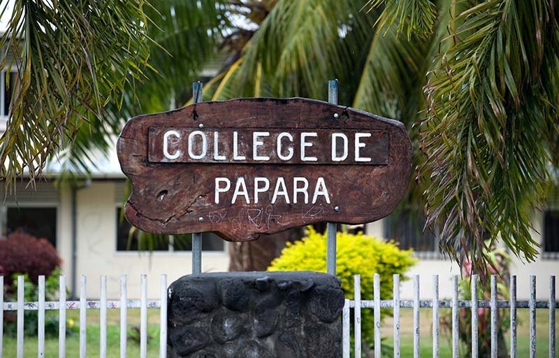 L'ancien panneau du collège : une planche de bois sombre gravée « COLLEGE DE PAPARA » en lettres blanches, montée sur deux tiges métalliques au-dessus d'un socle de pierre, sous les palmes.